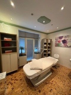 Klinischer Untersuchungsraum mit gepolsterter Behandlungsliege, Schränken, Regalen und floraler Wandkunst.