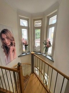 Treppenabsatz mit Erkerfenstern, Holzgeländer, rosa Blumen und einem großen Wandporträt einer Frau