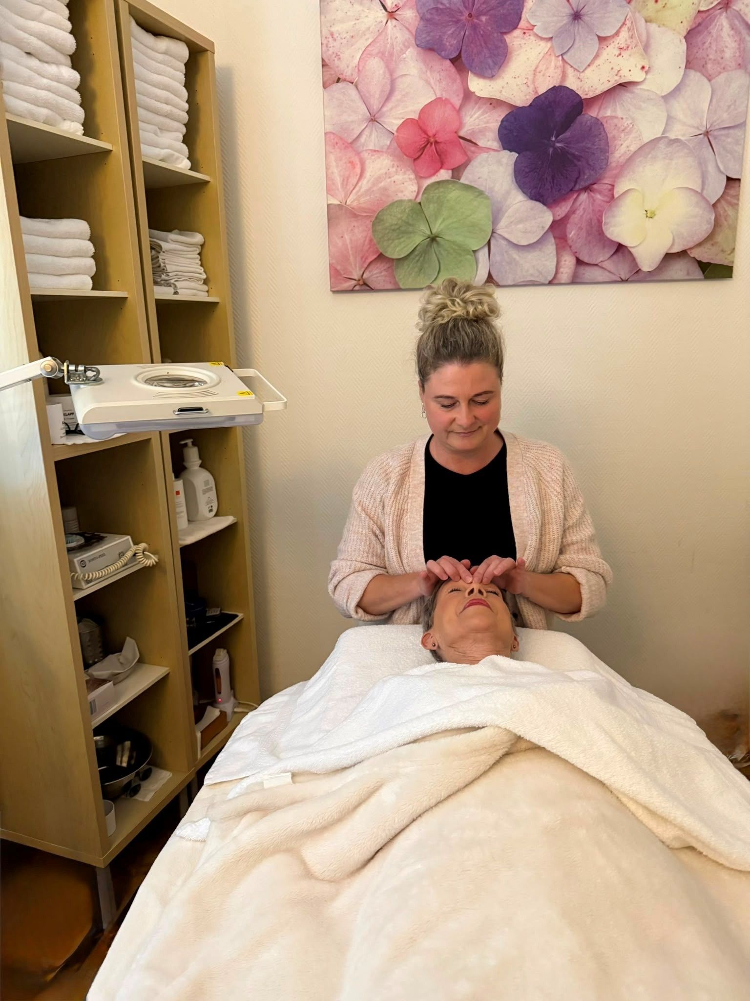 Eine Kosmetikerin gibt einer Kundin in einem Spa-Raum mit Handtüchern und floraler Wanddekoration eine Gesichtsmassage.