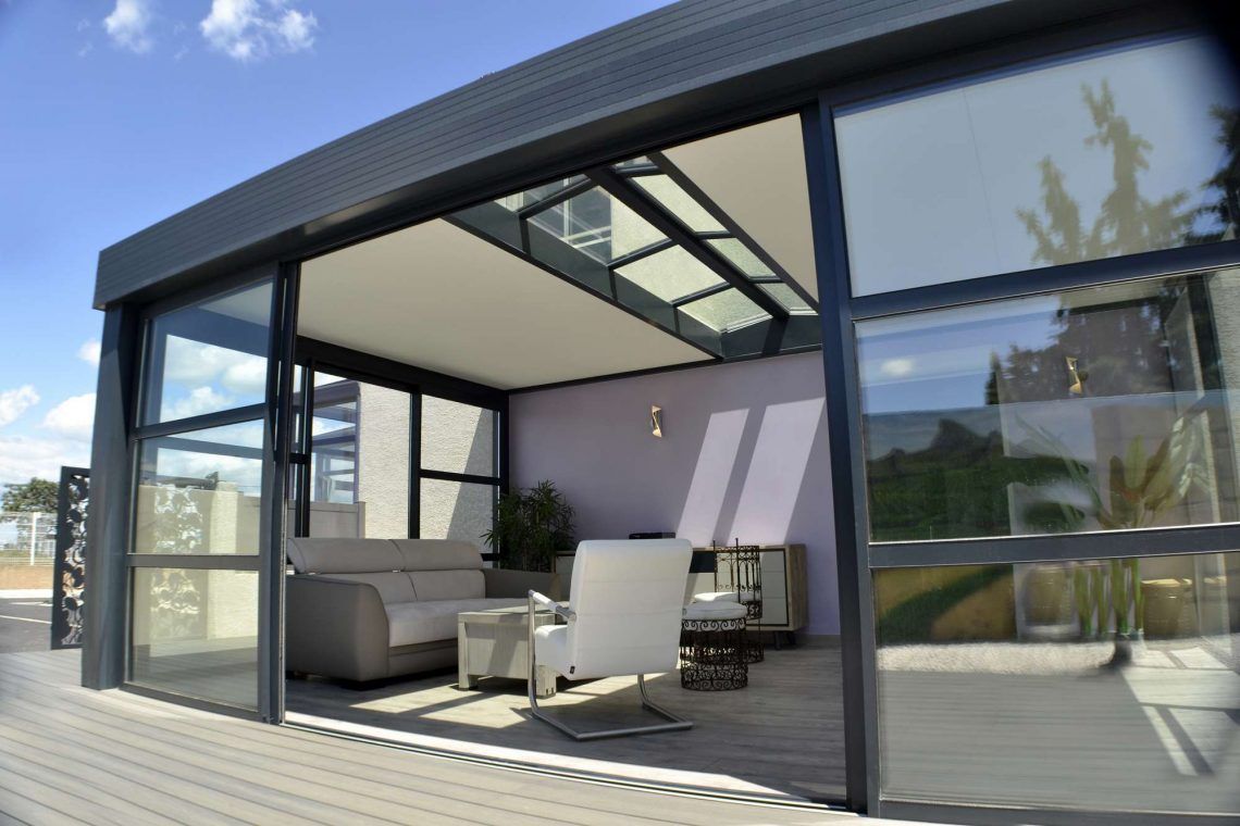 Véranda moderne avec cadre noir, chaises blanches et canapé gris sur une terrasse en bois sous un ciel bleu.