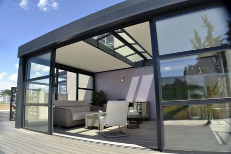 Véranda moderne avec cadre noir, chaises blanches et canapé gris sur une terrasse en bois sous un ciel bleu.