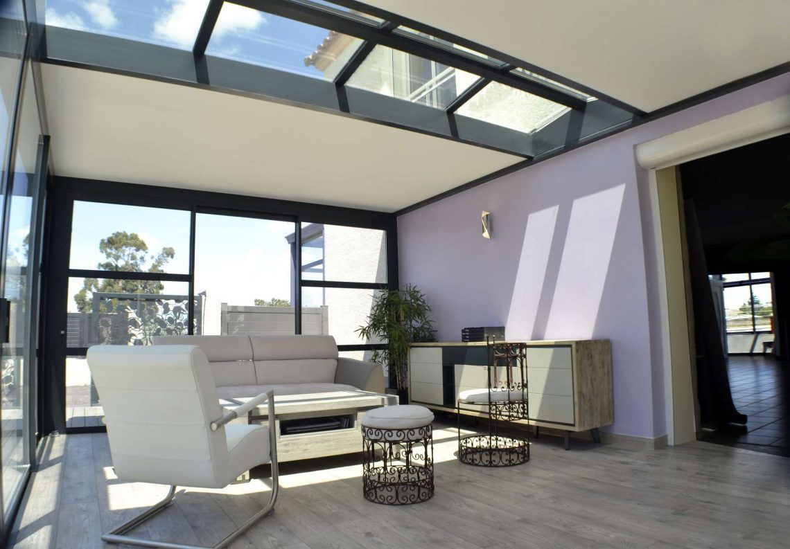 Salon moderne avec mobilier blanc, puits de lumière au plafond et grandes fenêtres donnant sur les arbres.