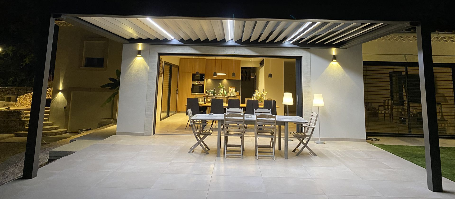 Terrasse extérieure avec table,chaises et lames de pergolas au-dessus, devant une maison éclairée la nuit.