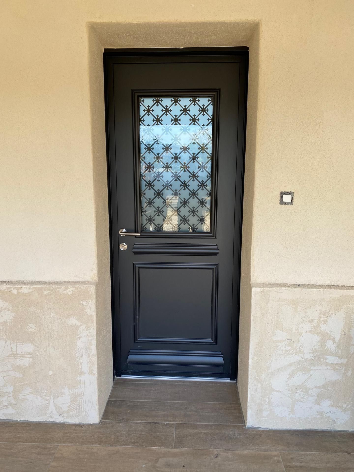 Porte d'entrée noire avec panneaux de verre dépoli, encastrée dans un mur extérieur beige.