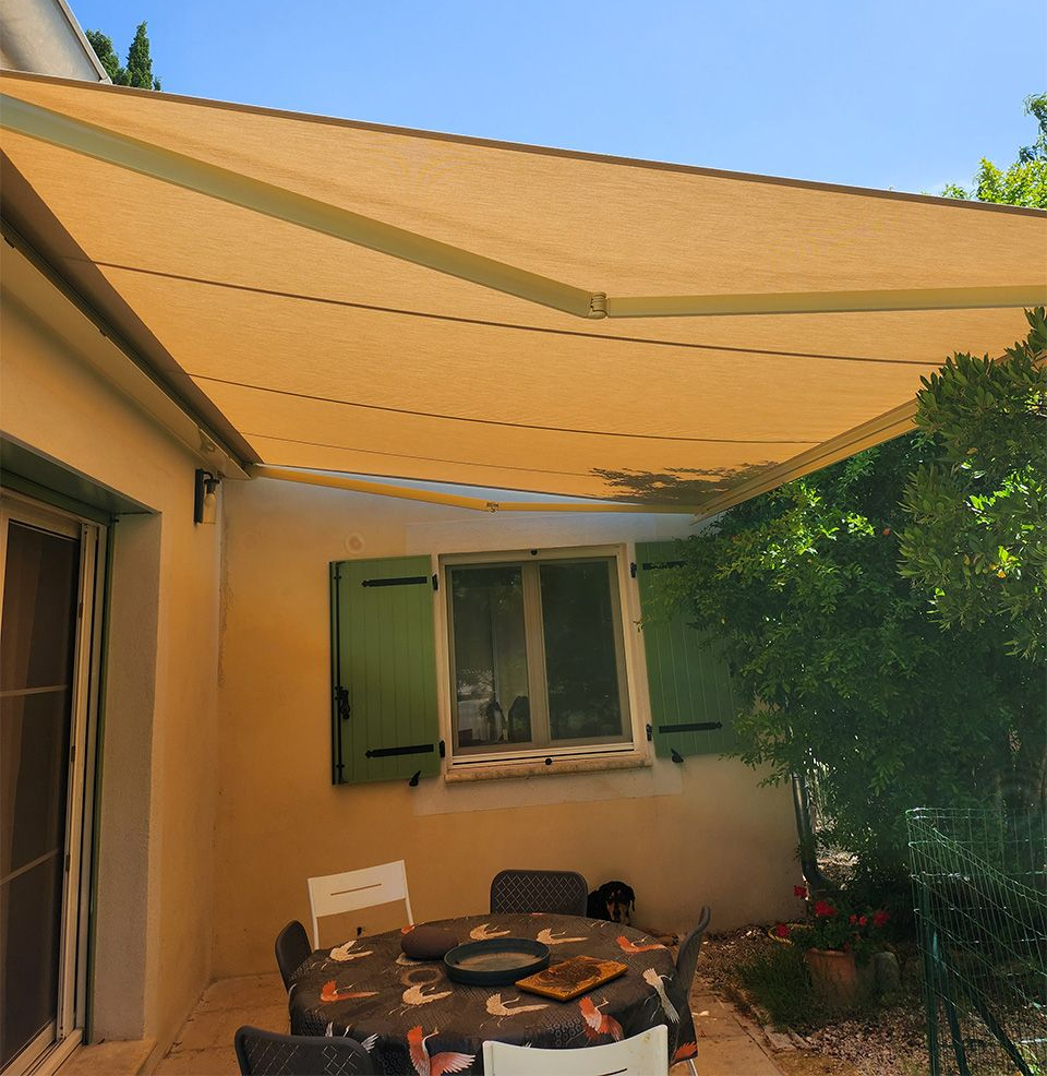 Terrasse avec store banne beige, table et chaises, et fenêtre de maison avec volets verts.