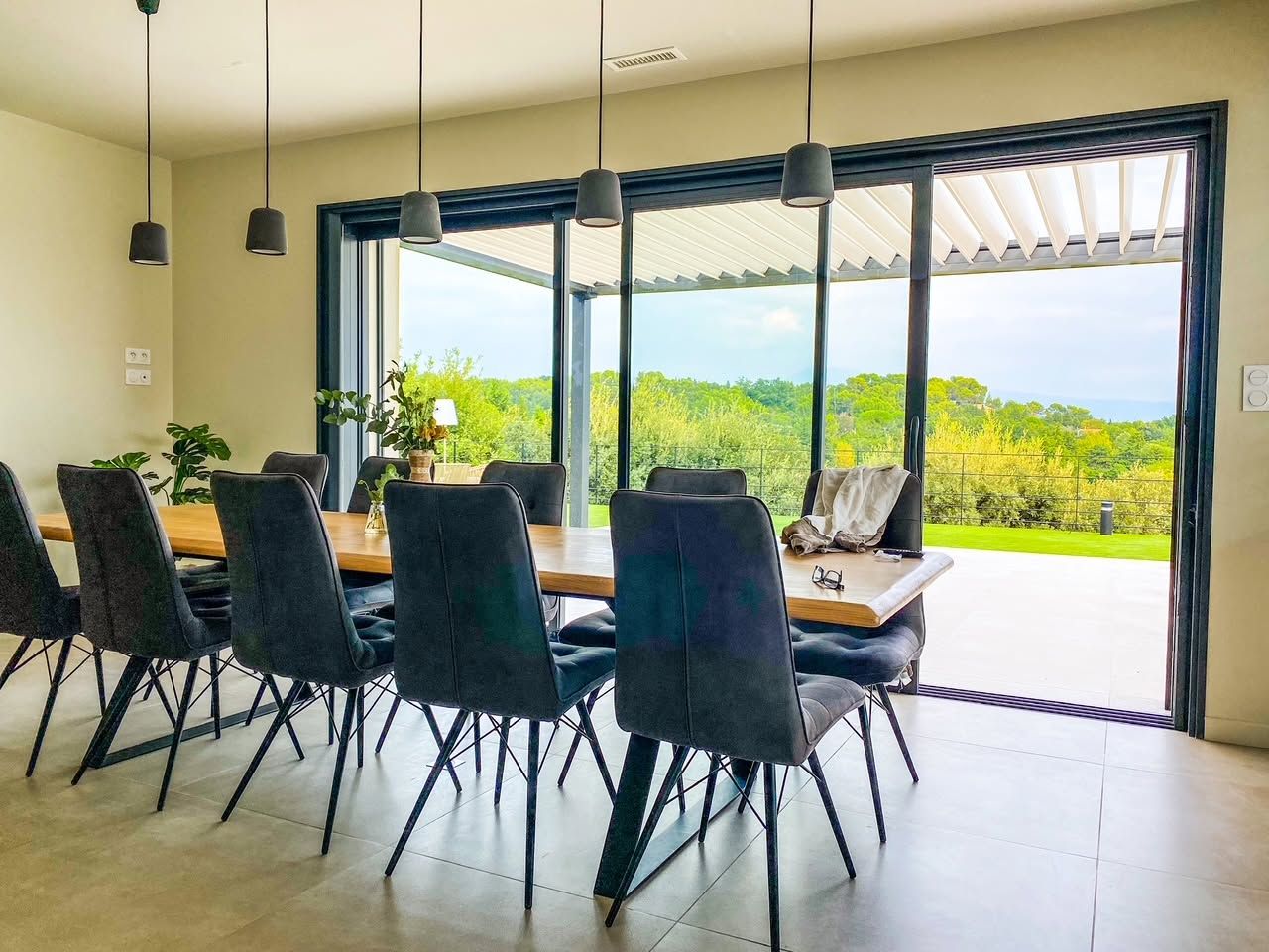 Salle à manger avec table et chaises dans une pièce avec baie vitrée donnant sur un paysage verdoyant et une terrasse.