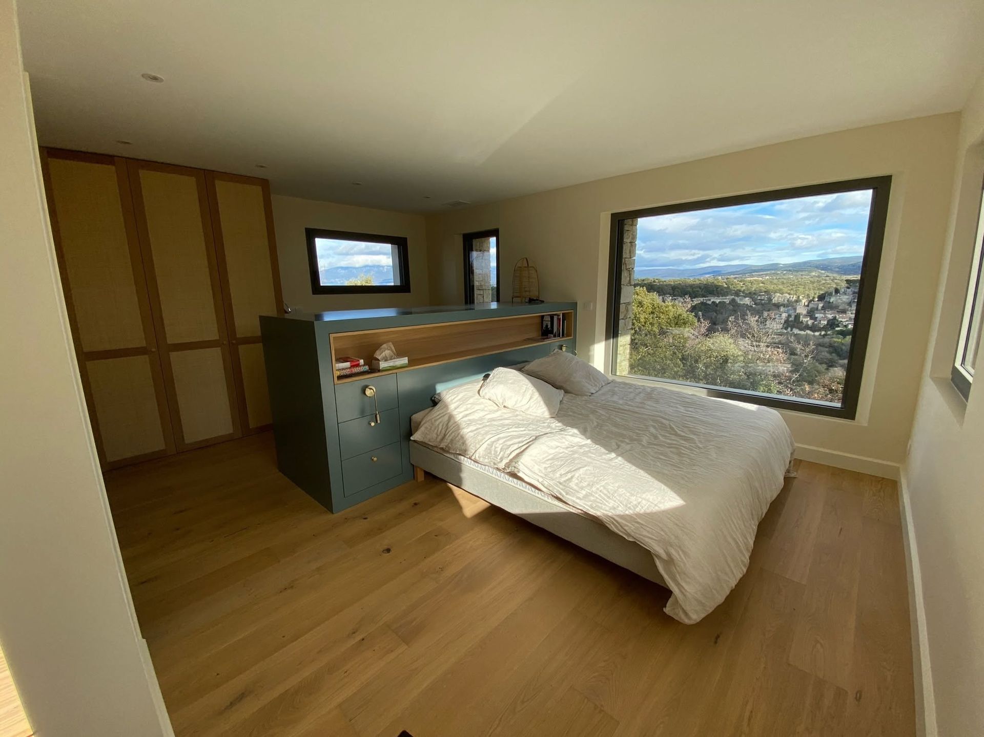 Chambre ensoleillée avec un lit, un parquet et une grande fenêtre donnant sur les collines.