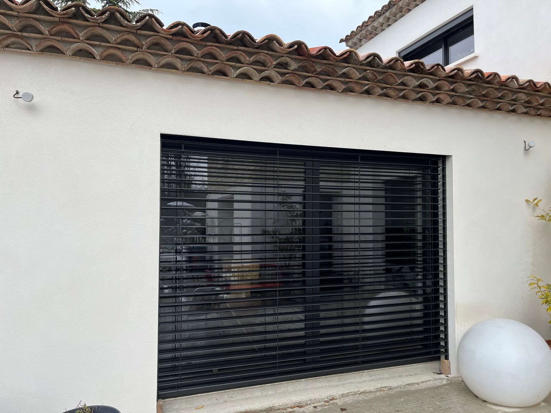 Mur d'une maison en stuc blanc avec une grande fenêtre à volets noirs et un objet sphérique blanc sur la terrasse.