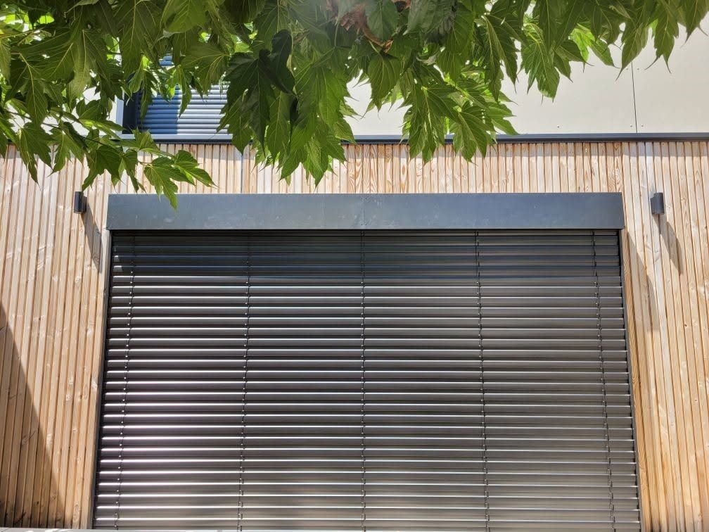 Store à lattes horizontales de type brise soleil orientable, sous des feuilles d'arbre vertes et façade de maison en bois.