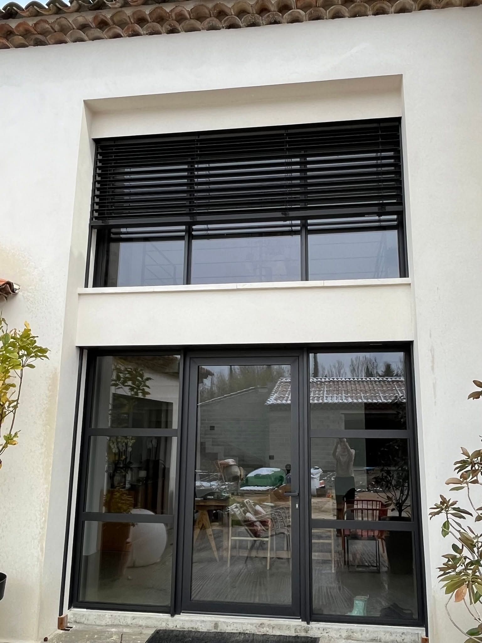 Façade moderne avec de grandes portes et fenêtres vitrées et brise-soleil orientable à lattes noires.