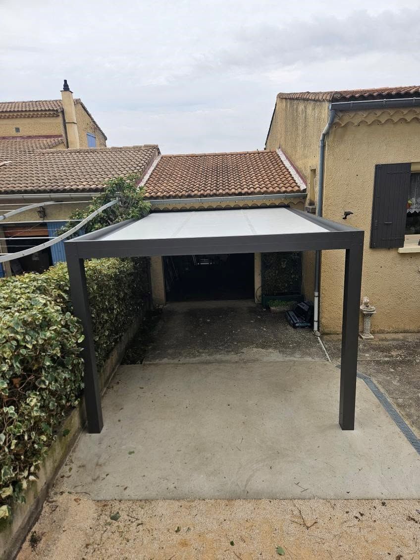 Abri voiture couvert dans une cour avec un toit gris, des poteaux noirs et des murs en pierre environnants.