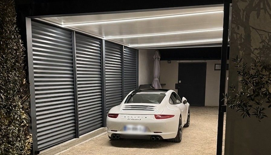Berline blanche garée sous un abri de voiture couvert, stores verticaux fermés, la nuit.