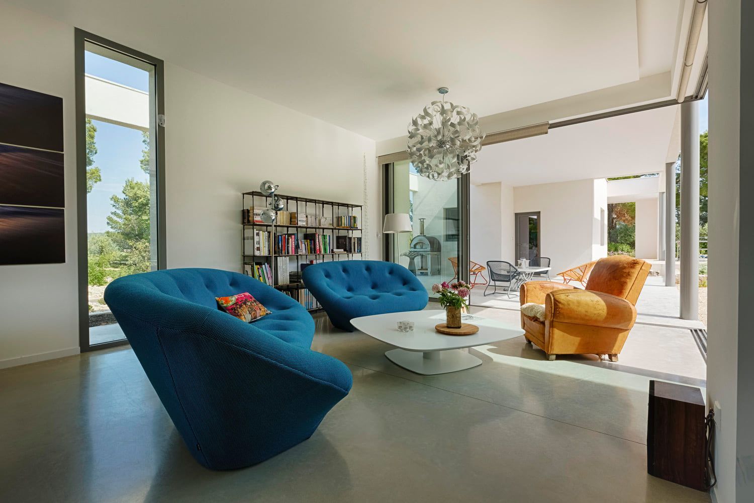 Salon moderne et lumineux avec des fauteuils bleus et jaunes, une table basse blanche et de grandes fenêtres.