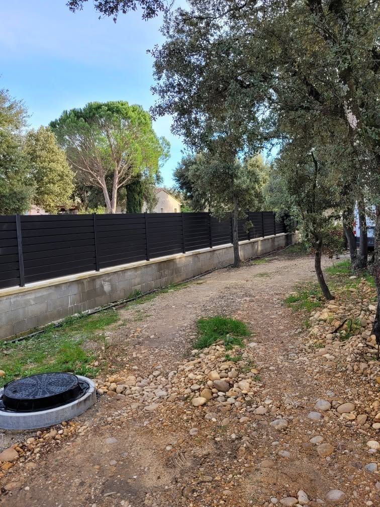 Chemin de terre en cours de travaux longeant une clôture noire et des arbres sous un ciel dégagé.