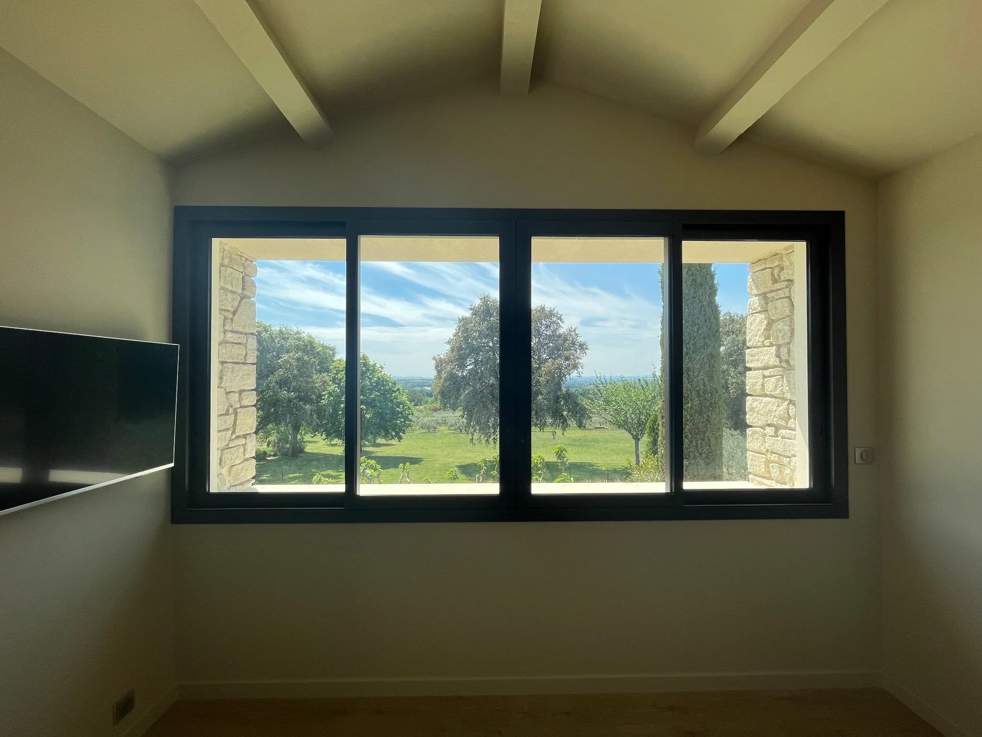 Pièce vide avec une grande fenêtre à cadre noir donnant sur un paysage verdoyant et un ciel bleu.