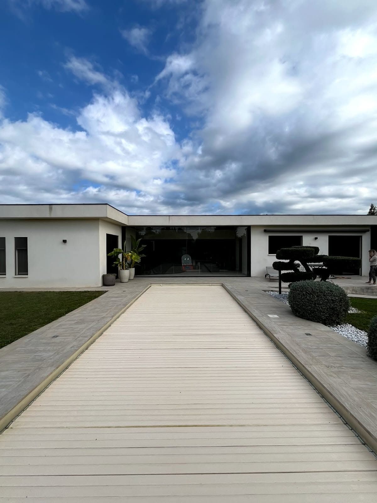 Maison blanche moderne avec une allée en bois menant à un patio sous un ciel nuageux.
