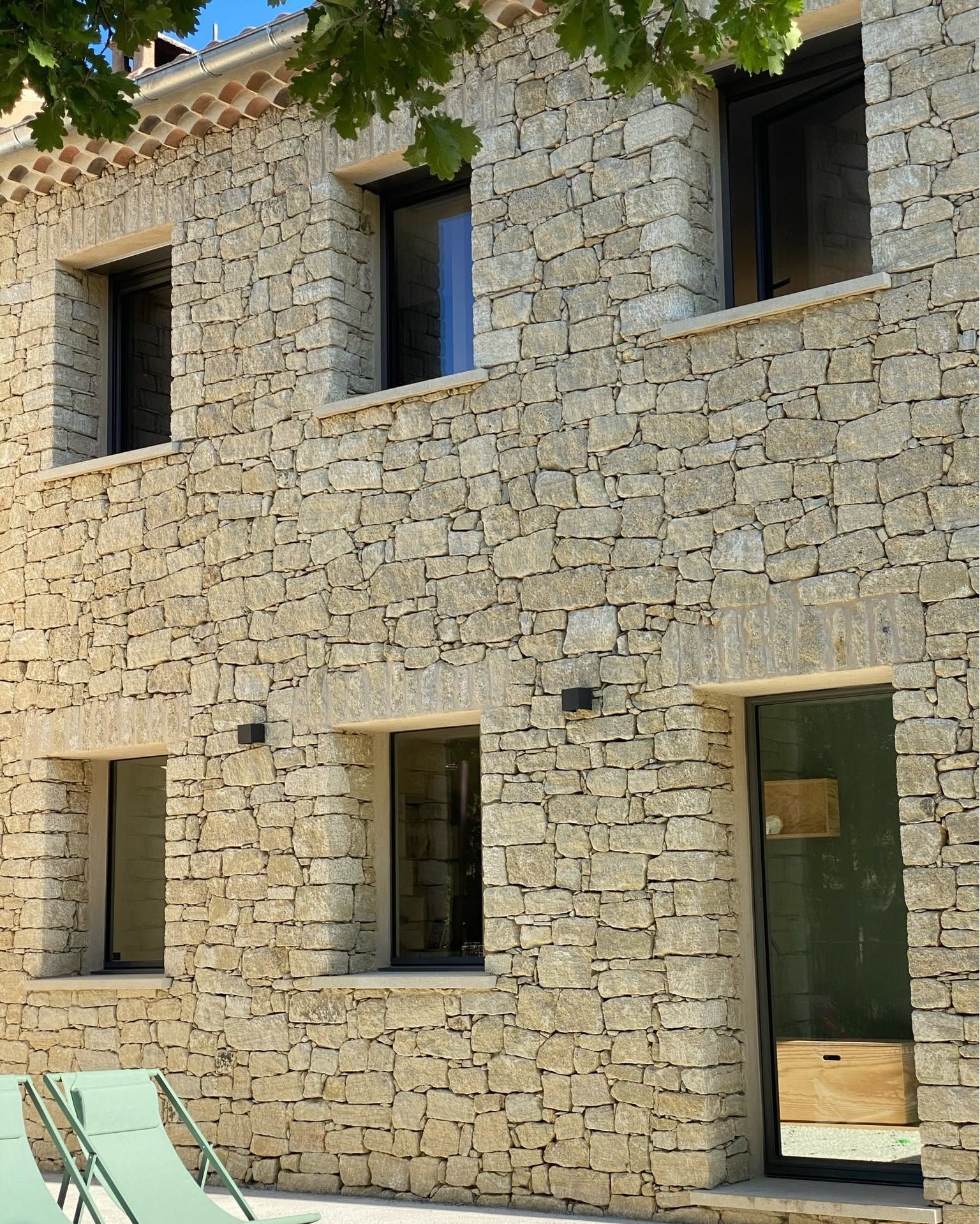 Façade d'une maison en pierre avec des fenêtres en aluminium, une porte, vue depuis un patio avec des chaises.