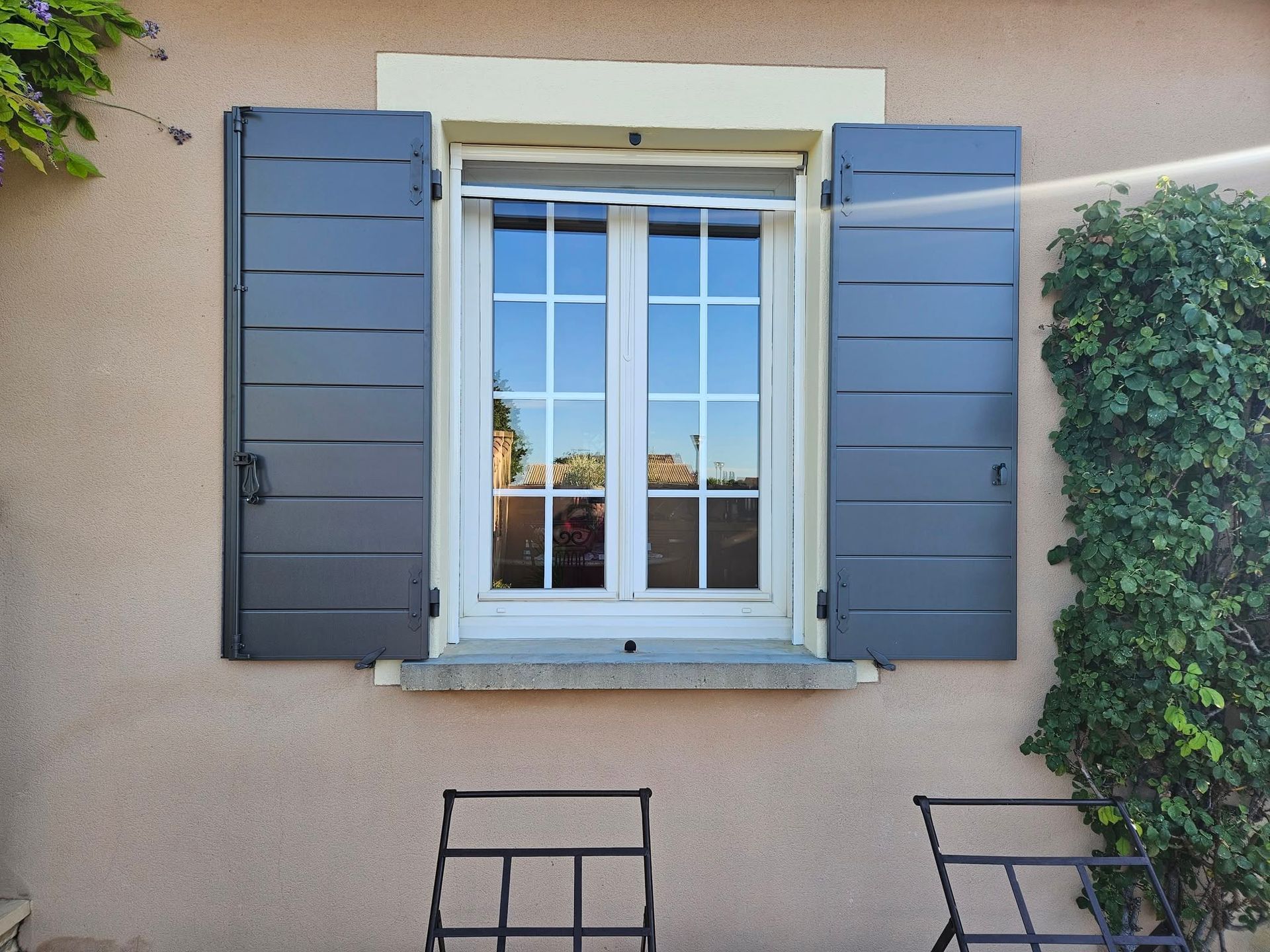 Fenêtre à cadre blanc et volets bleu foncé sur un mur beige, avec un arbuste en pot à côté.