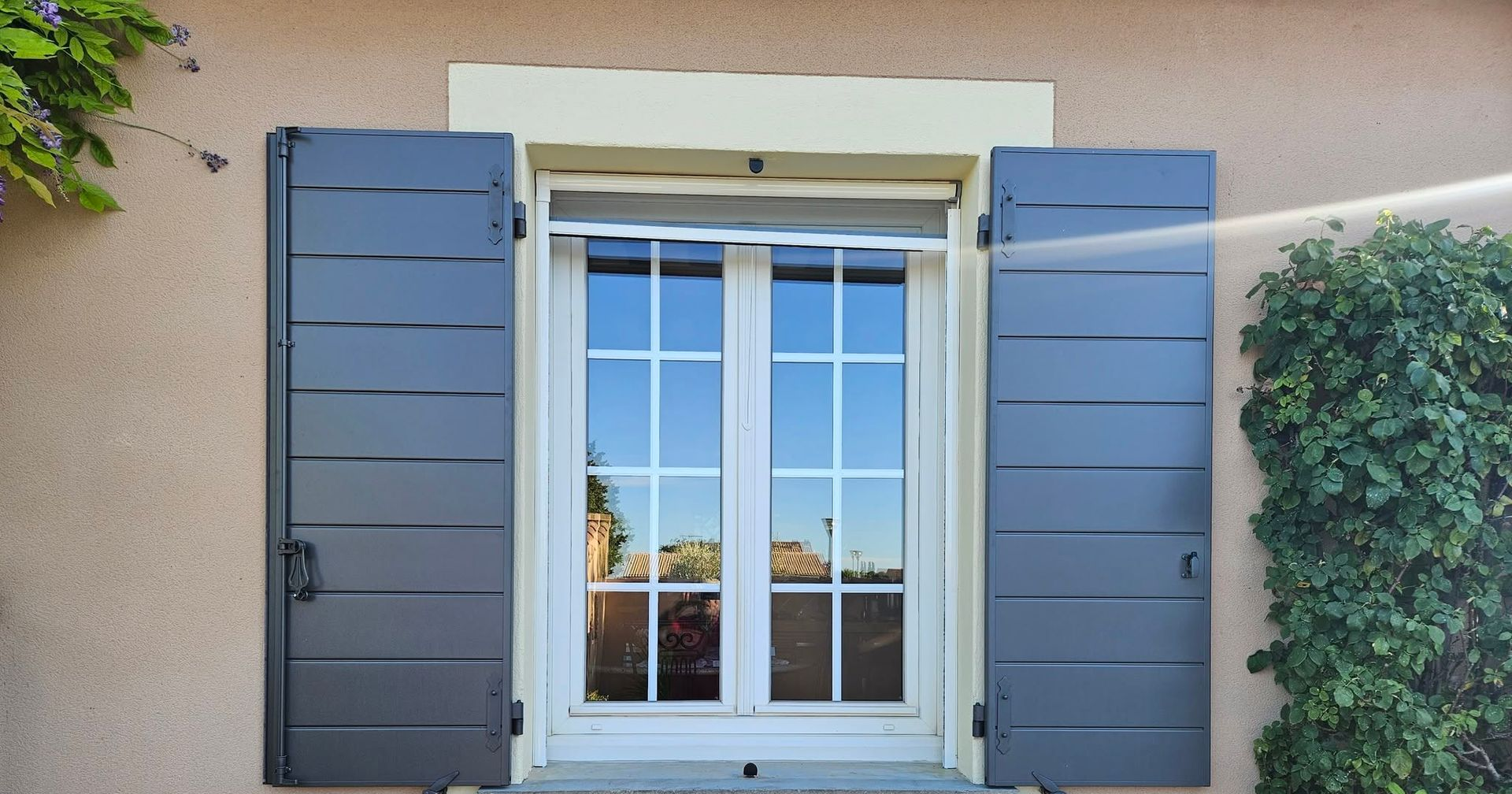 Fenêtre blanche avec moustiquaire enroulée et volets gris-bleu, sur une façade de maison beige.