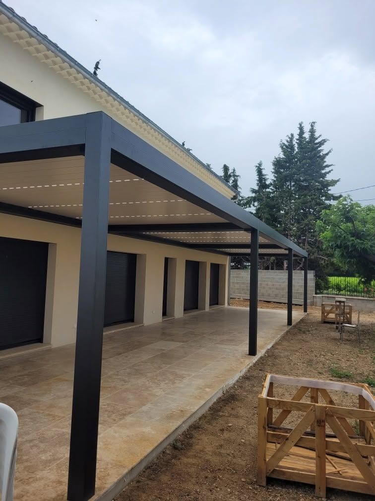 Patio de maison moderne avec colonnes noires, carreaux beiges et caisses en bois dans une cour en gravier.