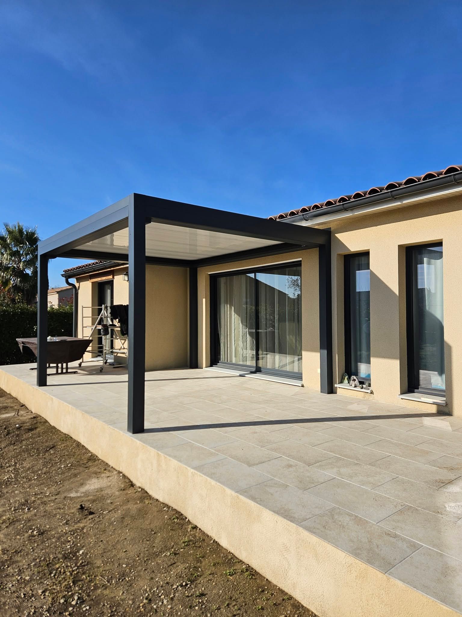 Patio d'une maison avec une pergola couverte, des portes coulissantes en verre et une terrasse pavée sous un ciel bleu.