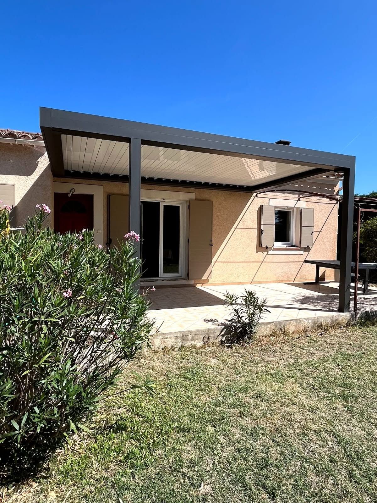 Maison de plain-pied de couleur beige avec un auvent noir à l'avant et un petit porche, vue depuis la cour.