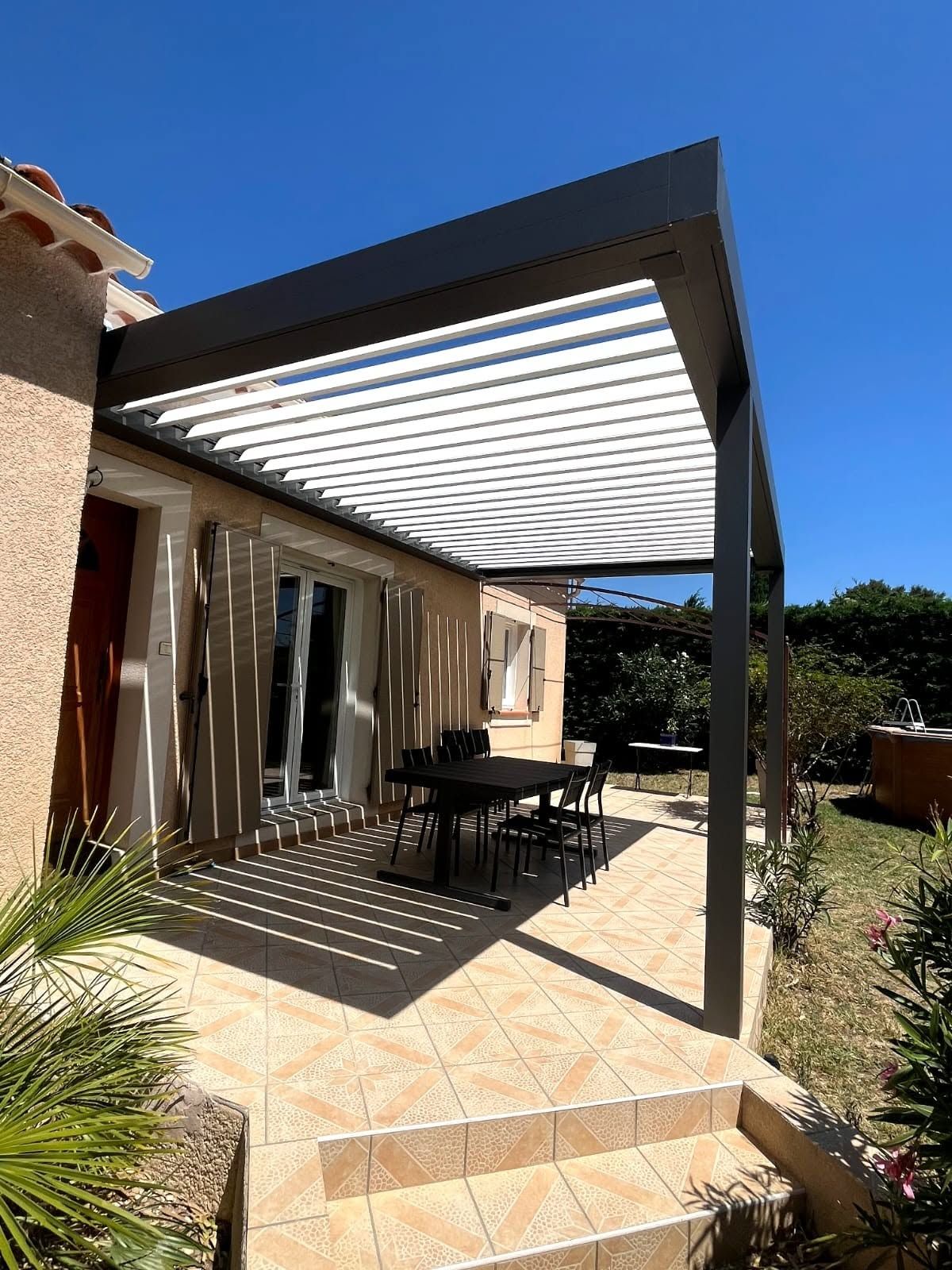 Terrasse avec pergola à lames orientables, table à manger extérieure devant une baie vitrée.