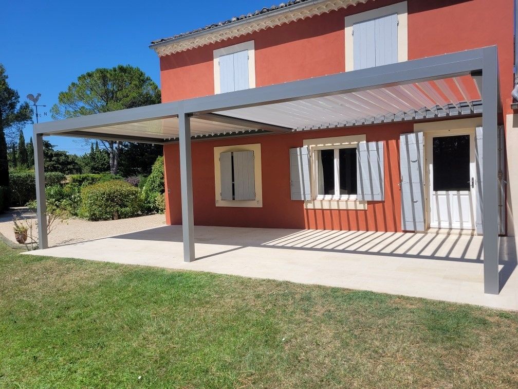 Maison rouge moderne avec patio couvert et pergola grise au-dessus d'une terrasse en béton, à côté d'un jardin gazonné.