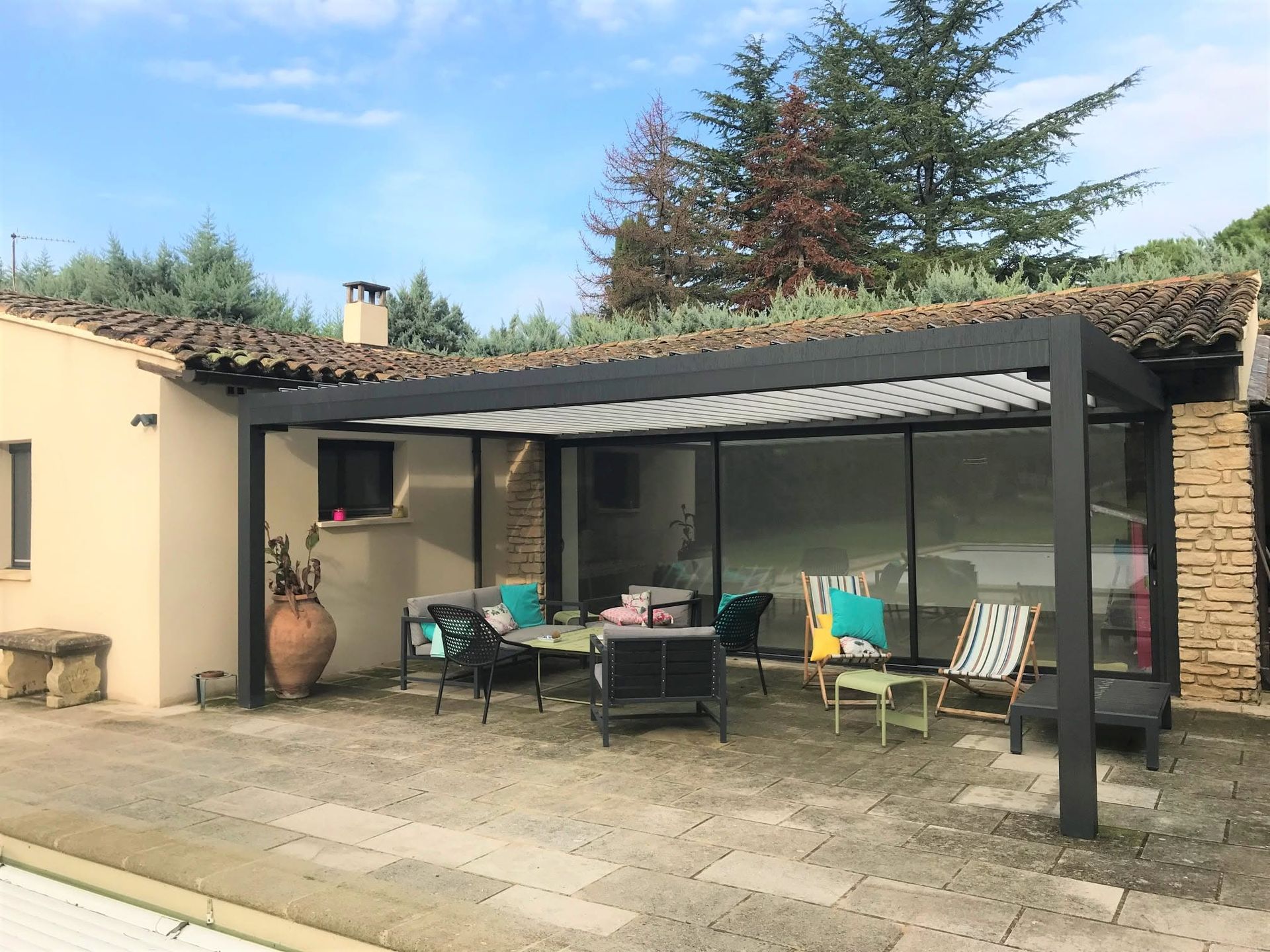 Terrasse avec sièges extérieurs sous une pergola, à côté d'une maison, avec des plantes en pot et des arbres au-delà.
