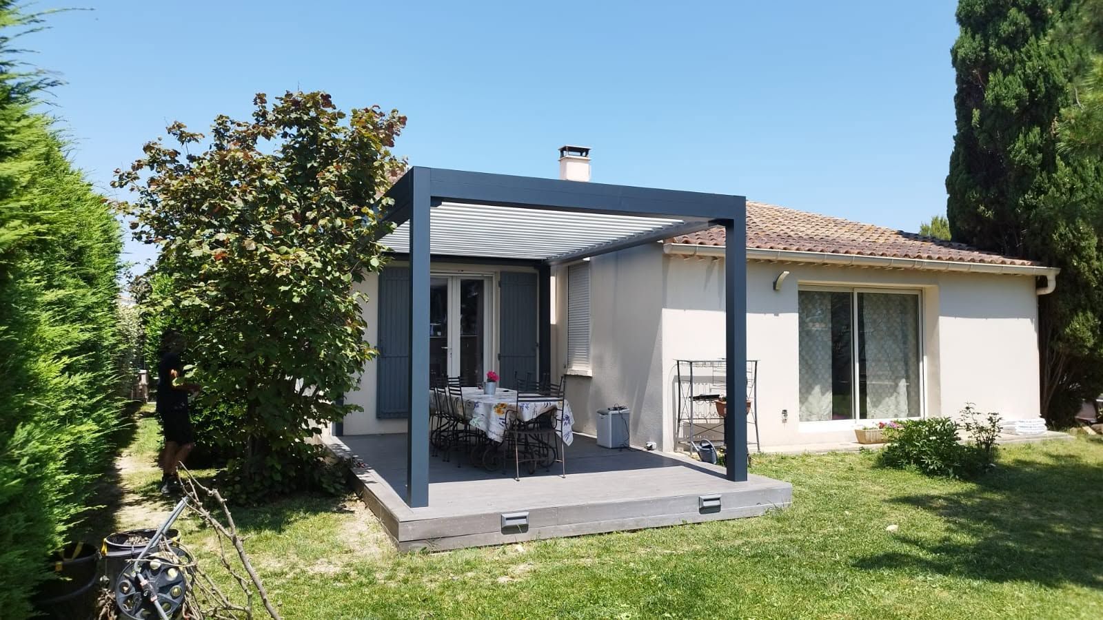 Patio d'une maison moderne avec pergola grise, table à manger extérieure et jardin gazonné sous un ciel bleu.