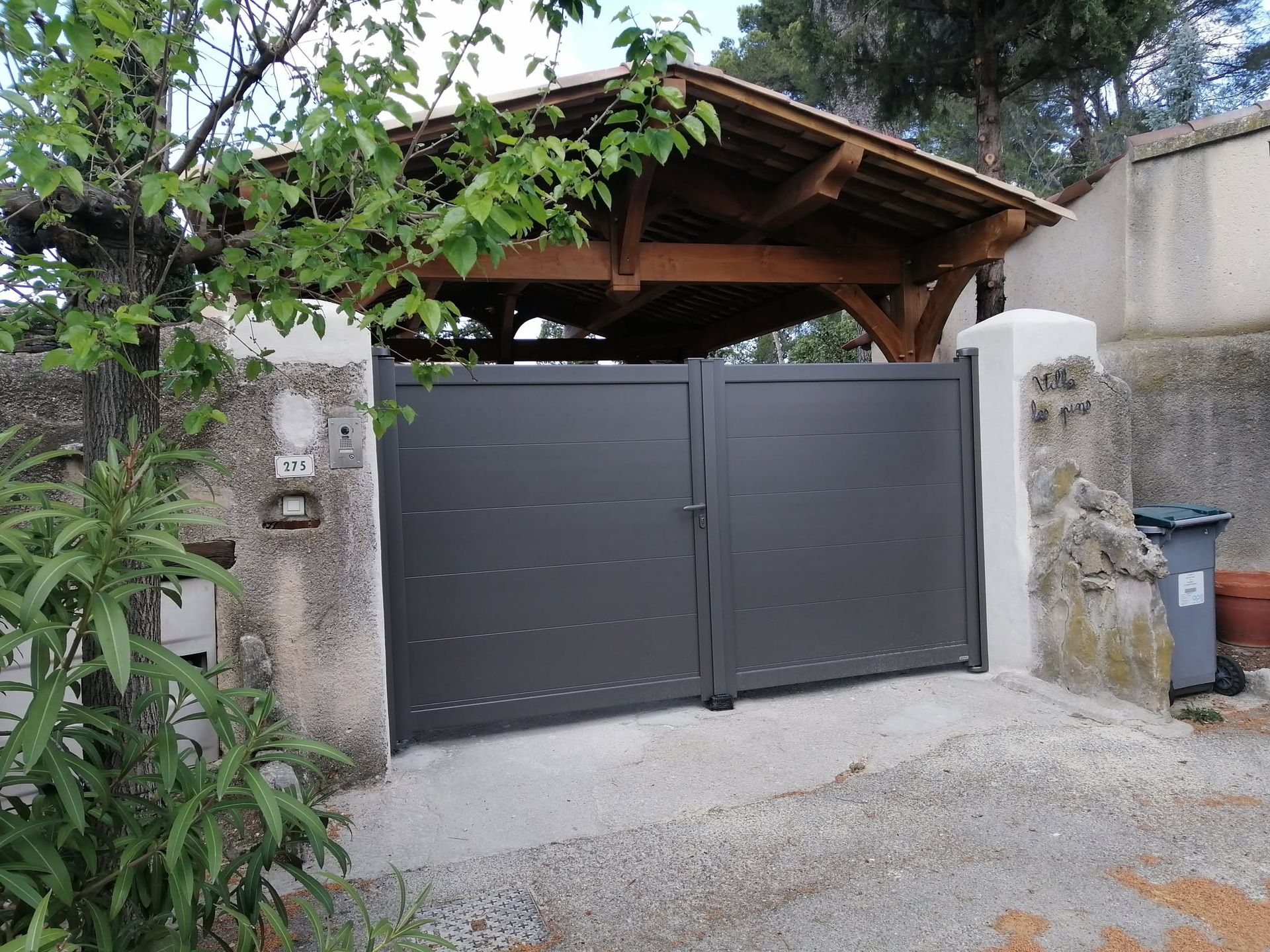 Portail double gris foncé qui marque l'entrée d'une allée et vue sur un abri en bois.