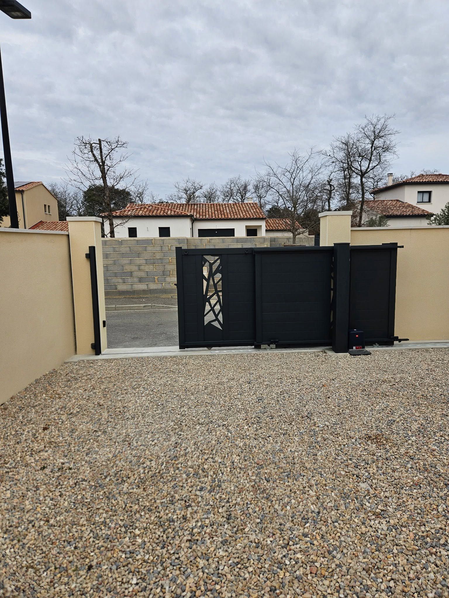 Portail en métal noir et mur beige dans une allée de gravier sous un ciel nuageux.