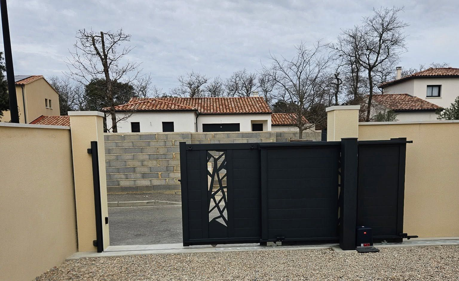 Portail coulissant noir avec mur beige. Mur en parpaing, maison et arbres dénudés en arrière-plan.