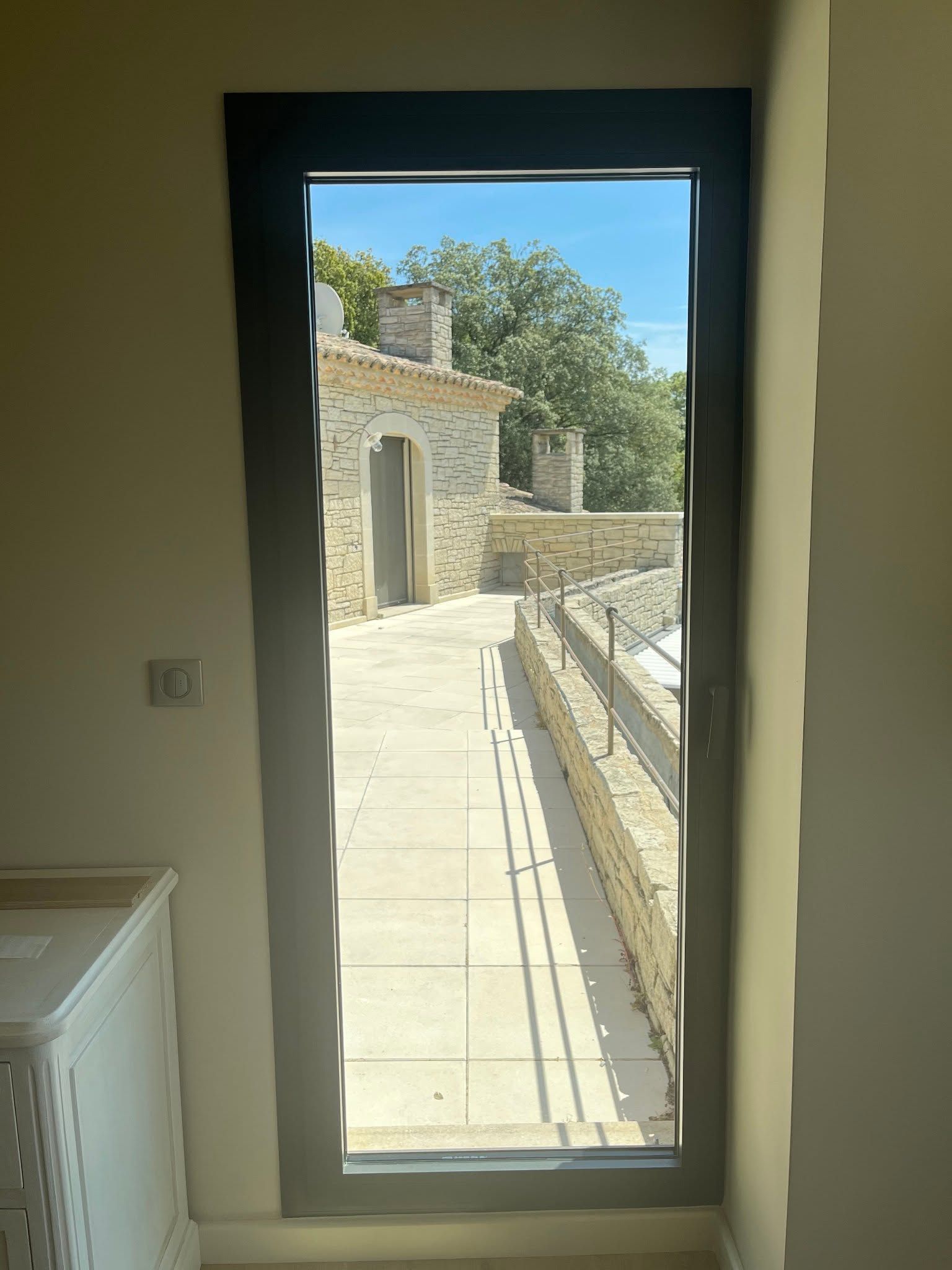 Porte vitrée donnant sur une terrasse en pierre ensoleillée et un bâtiment sous un ciel bleu.