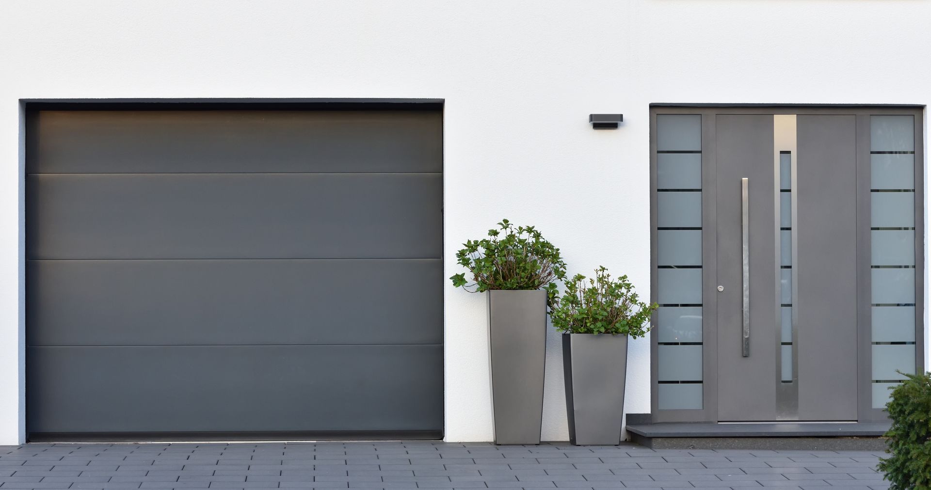 Porte de garage grise moderne et porte d'entrée assortie, plantes en pot sur un mur blanc.
