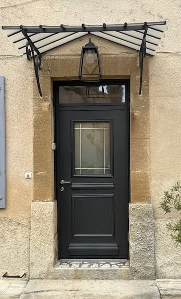Porte d'entrée noire avec lanterne sous un auvent métallique dans une entrée en pierre.