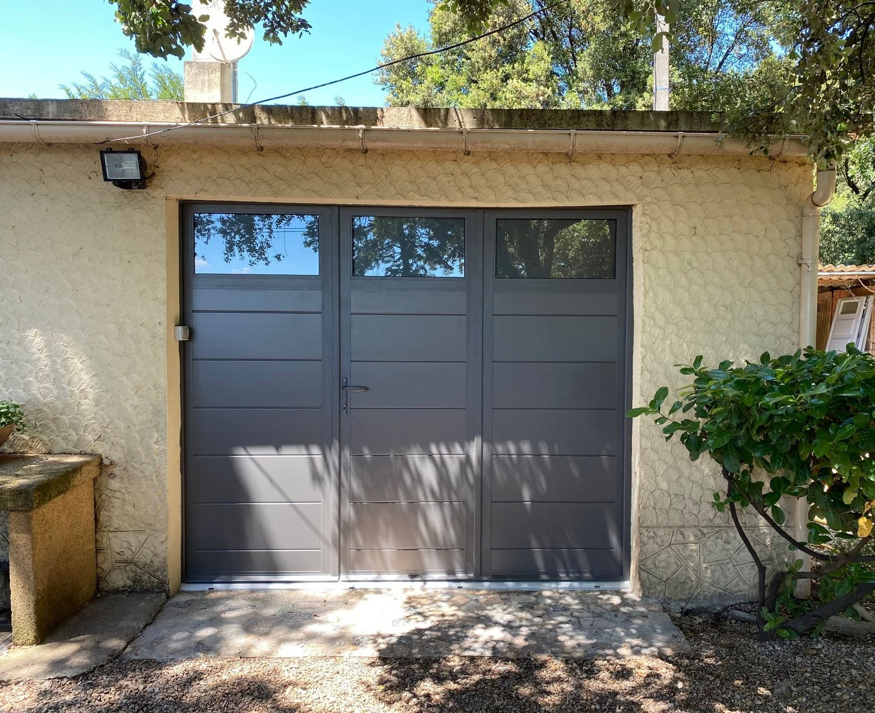 Porte de garage double grise sur une maison beige, avec des arbres et des arbustes devant.