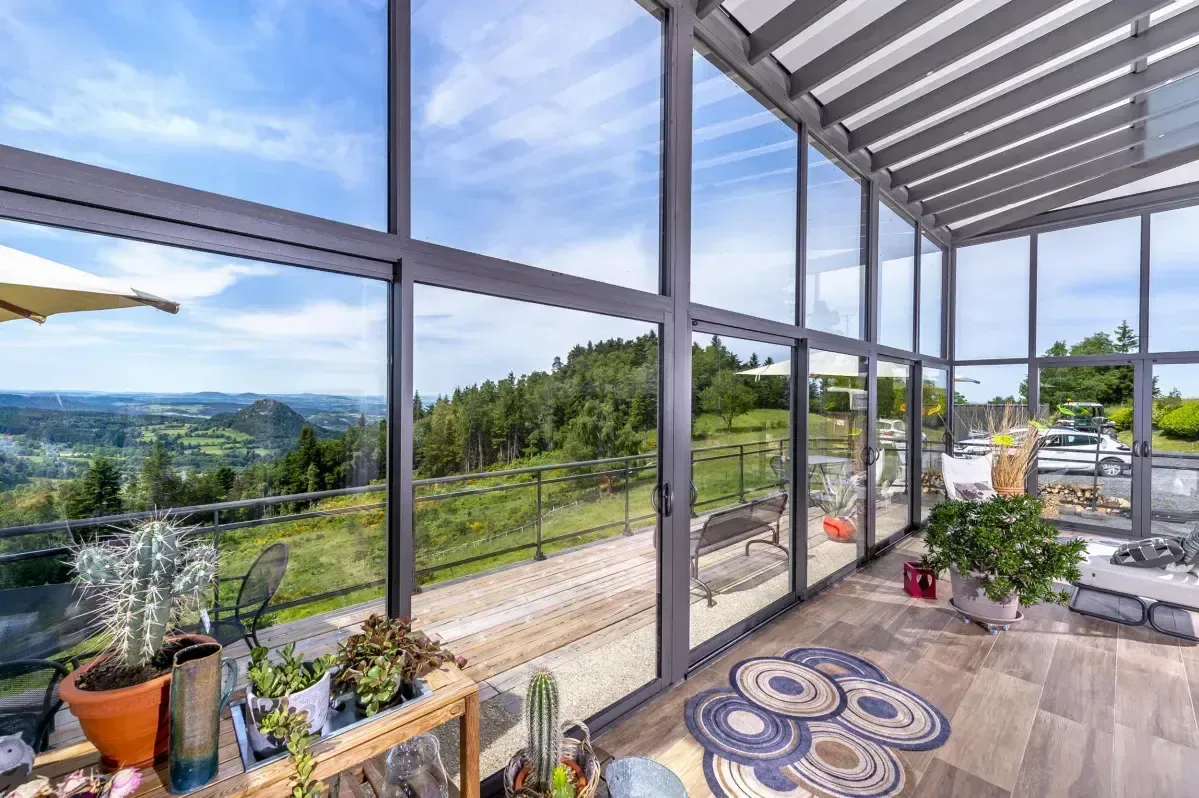 Véranda ensoleillée avec plantes en pot, sol avec motifs et larges fenêtres donnant sur un paysage verdoyant.