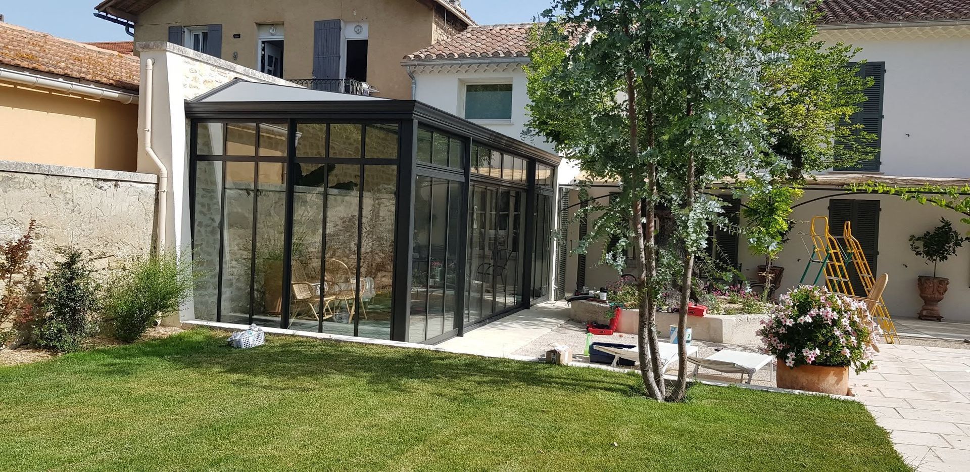Terrasse arrière avec véranda vitrée, pelouse, arbres et allée pavée longeant une maison.