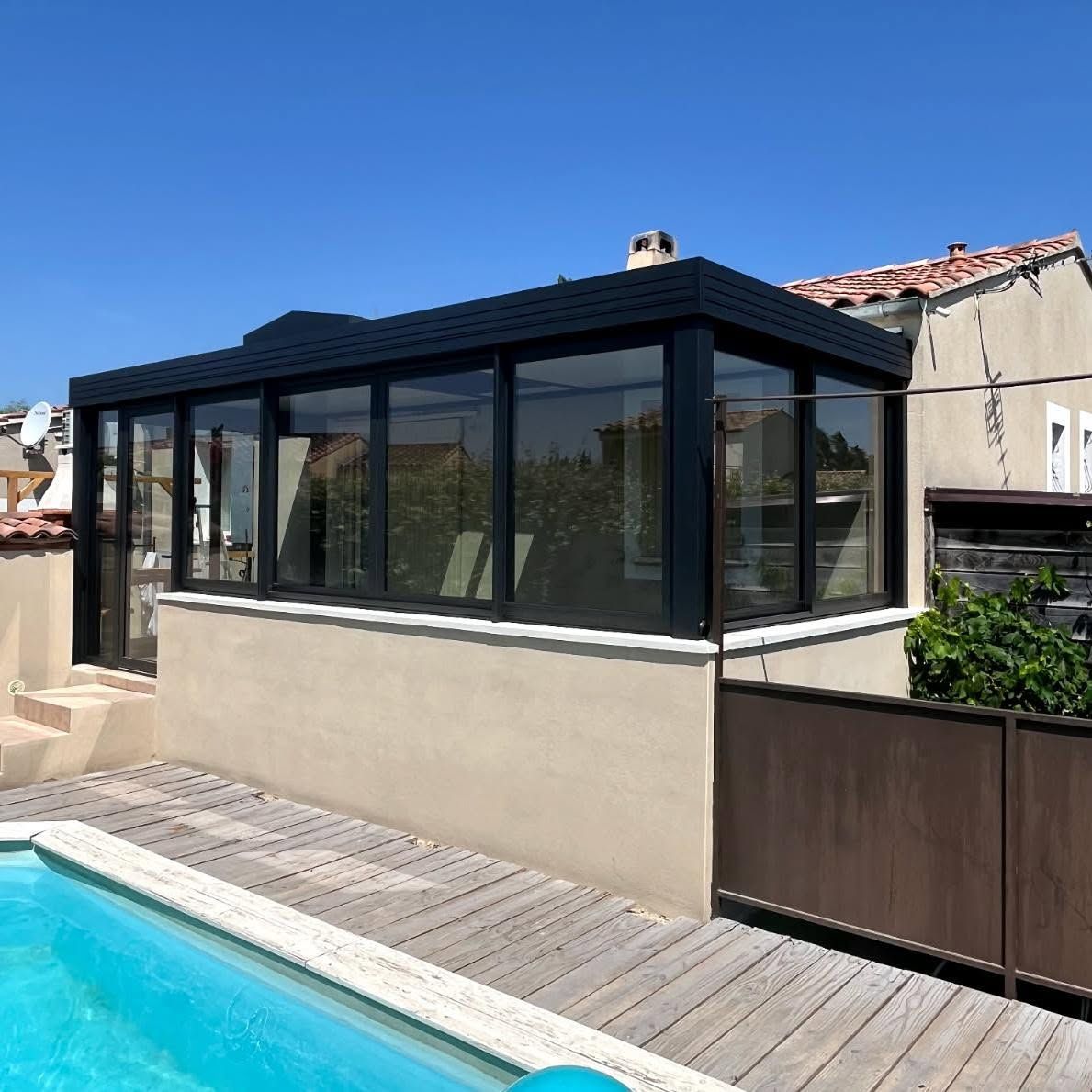 Véranda fermée attenante à une piscine, avec fenêtres à cadre noir et toit sombre.