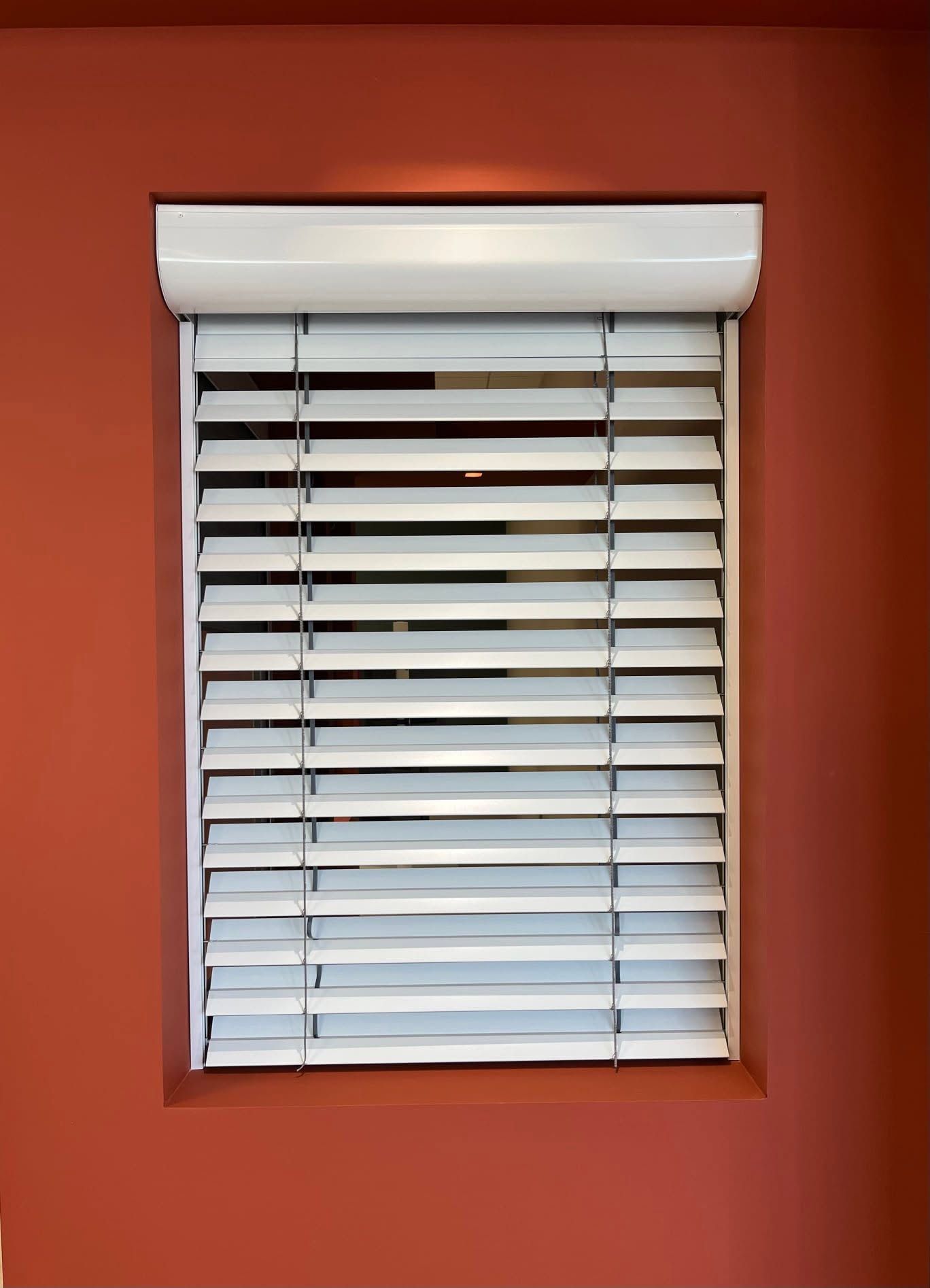 Volet roulant et brise-soleil blancs sur un mur rouge orangé.