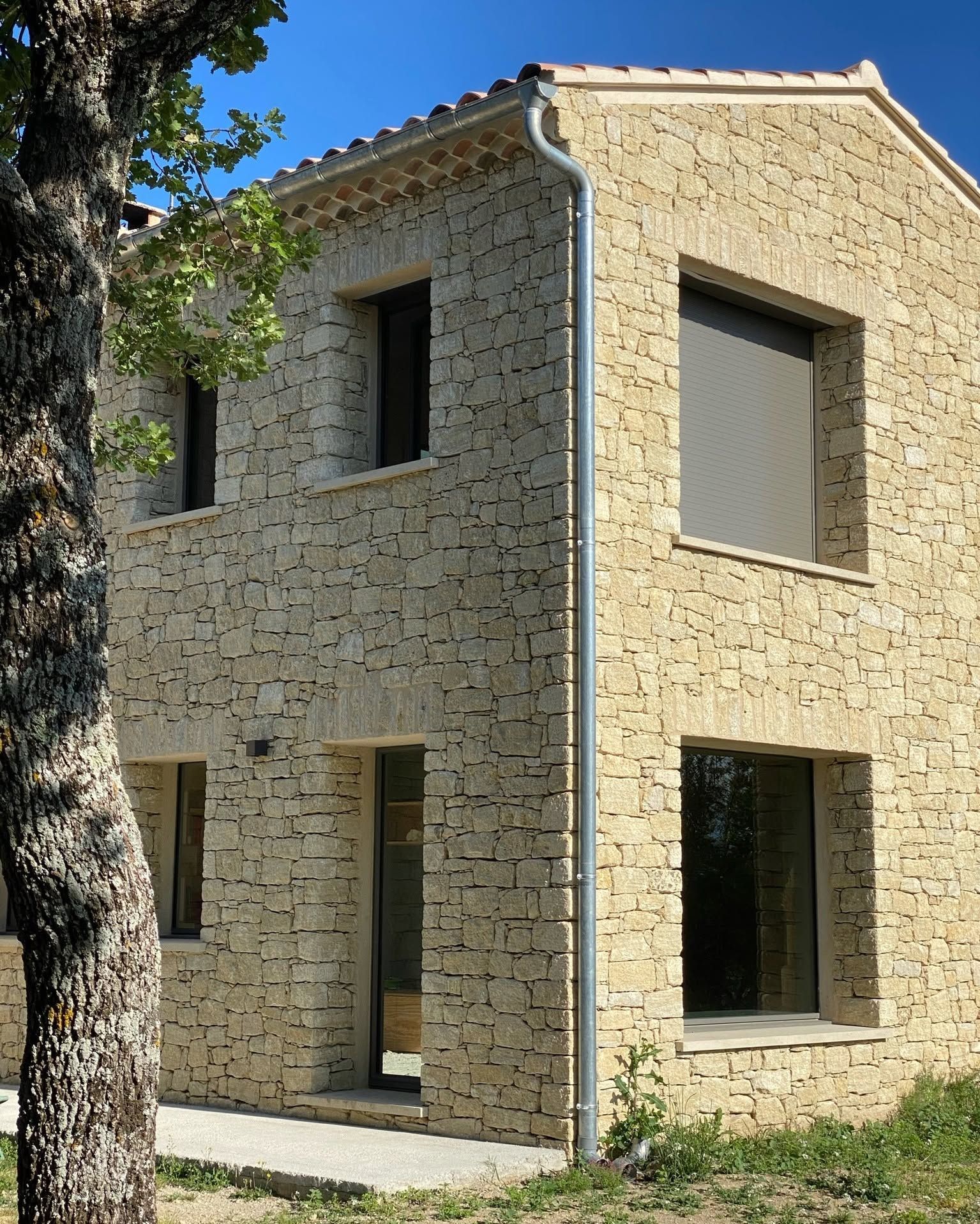 Maison en pierre de deux étages avec des boiseries grises et de grandes fenêtres, située à côté d'un arbre et d'une pelouse.