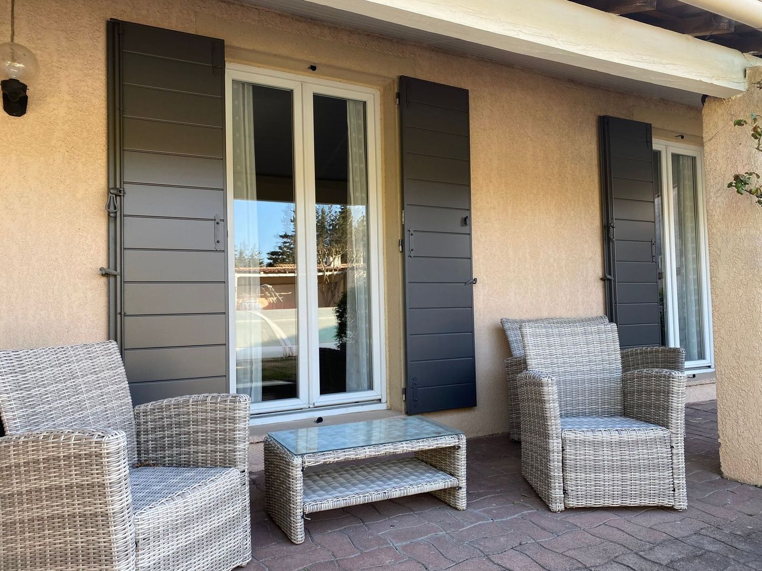 Deux chaises en osier et une petite table sur une terrasse beige avec des volets foncés et une porte vitrée.