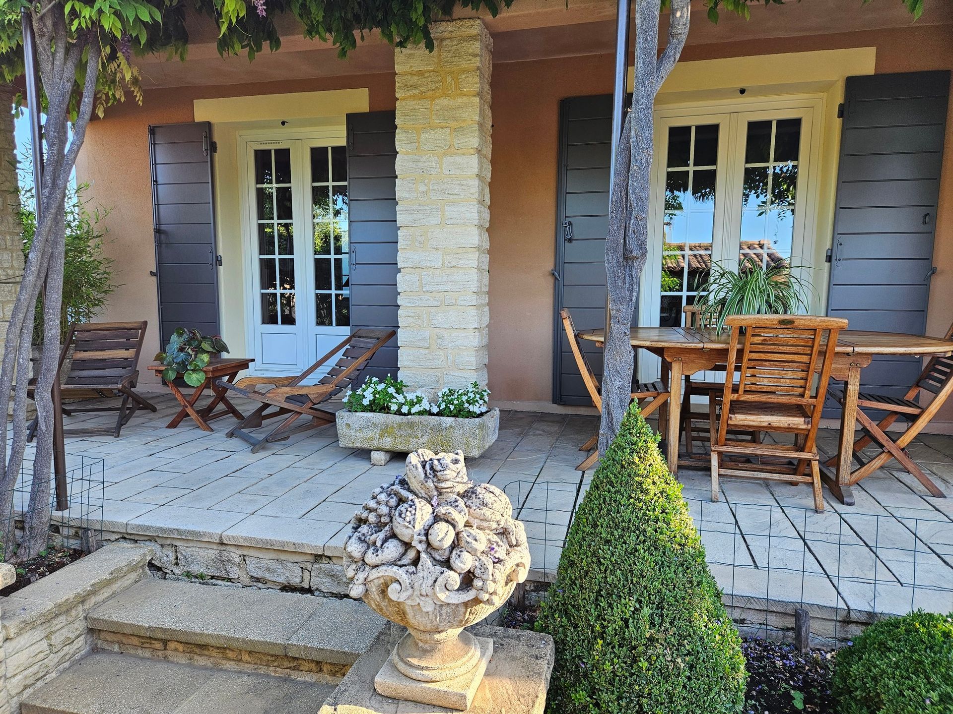 Une maison rustique avec pilier en pierre, portes-fenêtres et volets, mobilier de jardin en bois et une plante en pot.