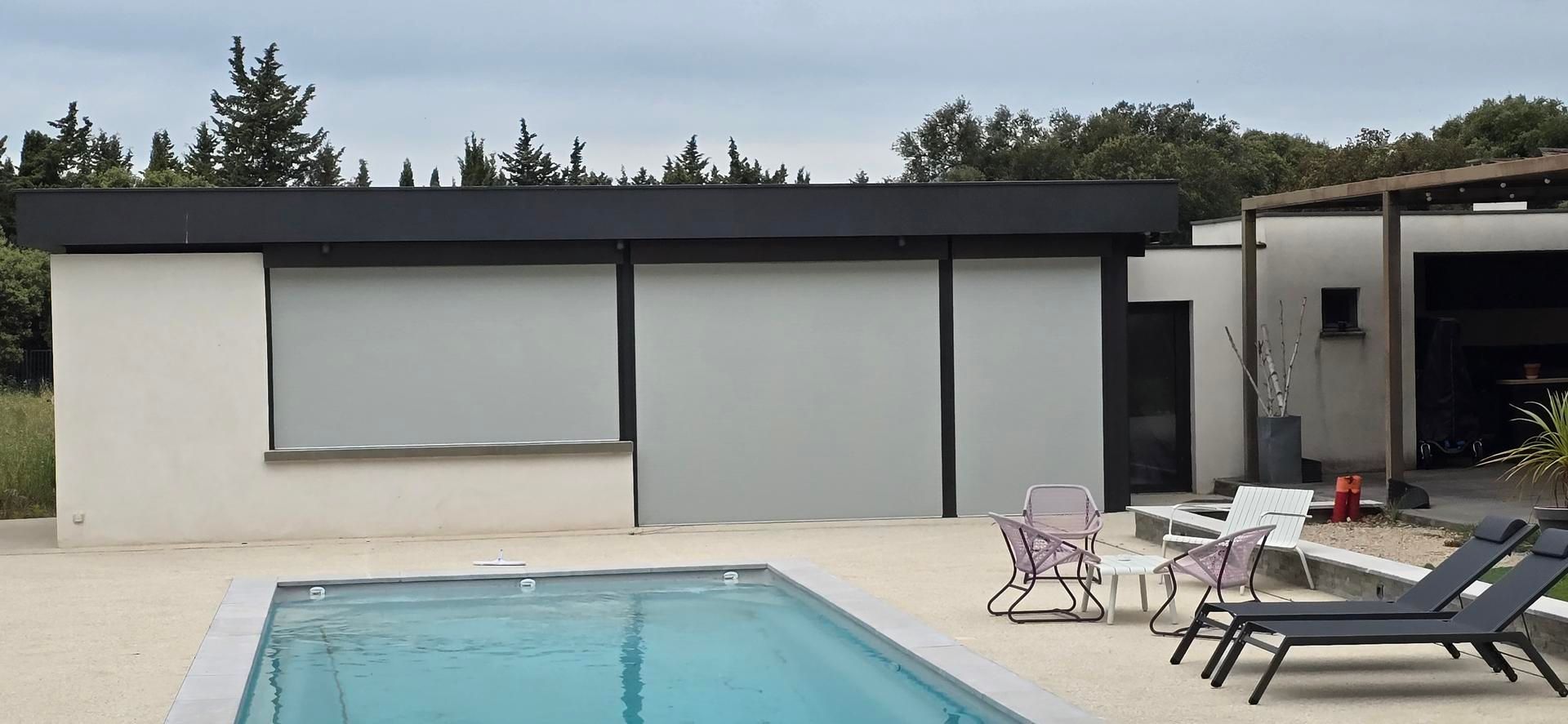 Patio d'une maison moderne avec piscine, chaises longues et grandes fenêtres protégées par des volets roulants.