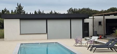 Patio d'une maison moderne avec piscine, chaises longues et grandes fenêtres protégées par des volets roulants.
