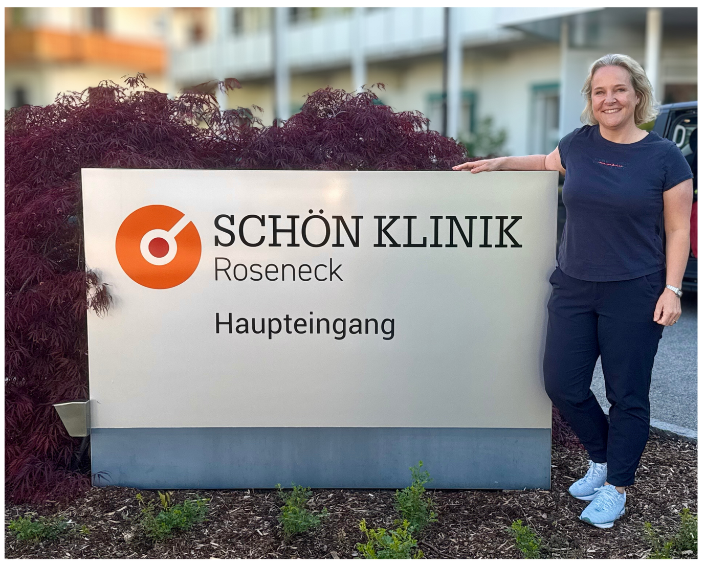 Julia Reher steht neben dem Schild der Schön Klinik Roseneck vor dem Klinikgebäude. 