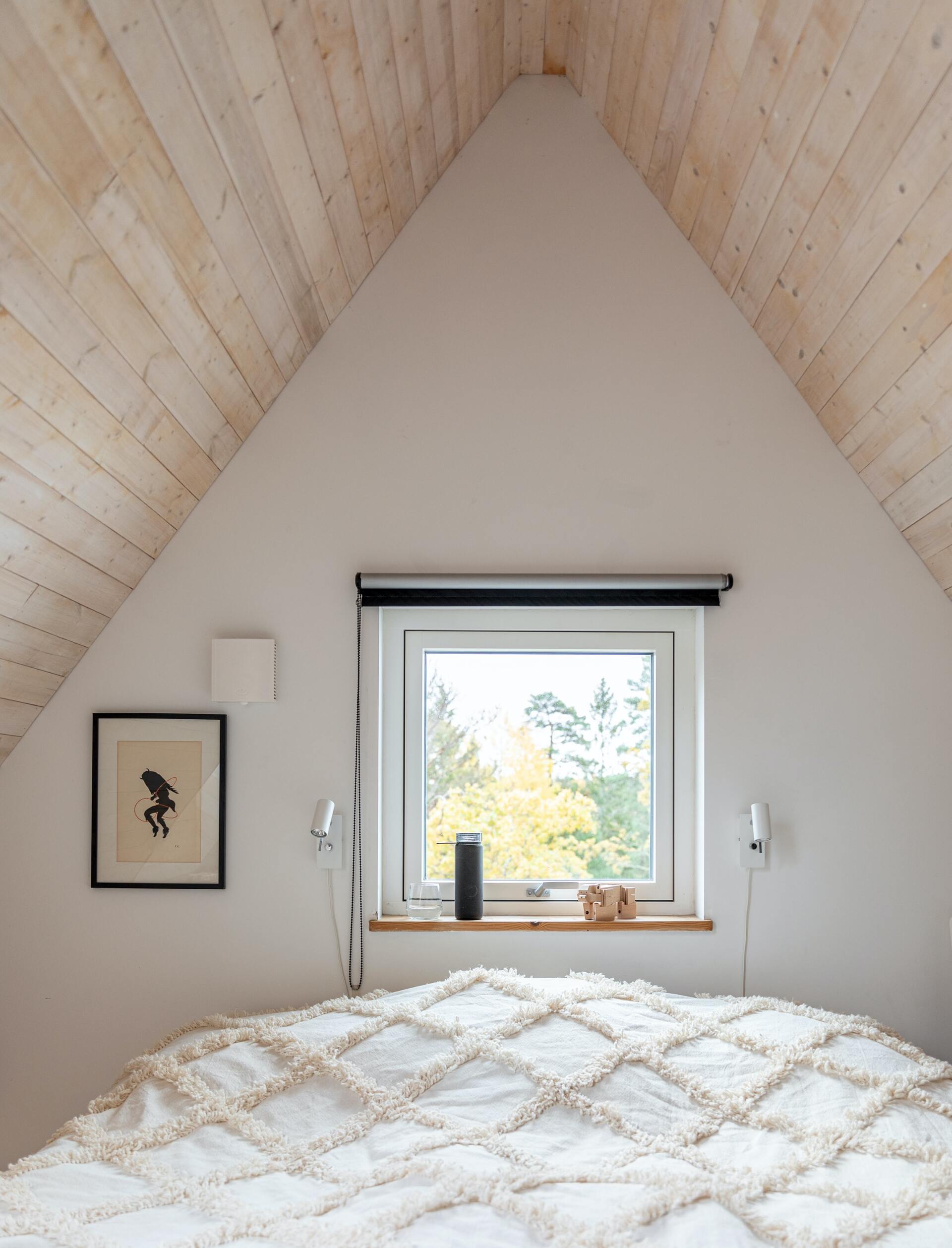 Chambre avec plafond en pente en bois blanc, fenêtre au-dessus du lit, œuvres d'art au mur.