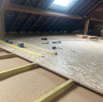 Grenier avec plancher en bois partiellement installé et poutres apparentes, lumière naturelle provenant d'un puits de lumière.