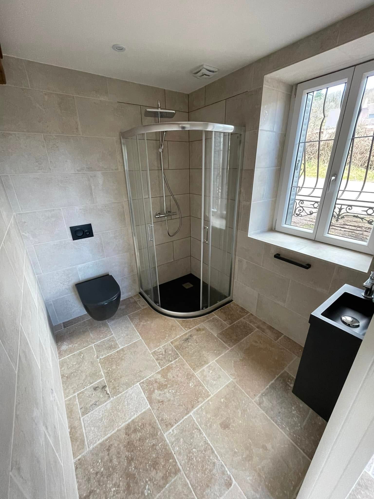 Salle de bains avec carrelage en pierre beige, toilettes noires, douche en verre, meuble-lavabo noir et fenêtre.